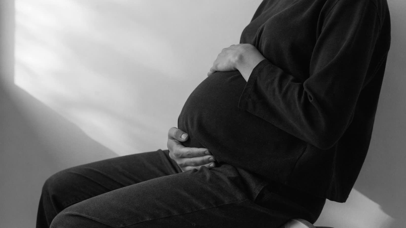 Gestante sentada de perfil, do ombro para baixo, com as mãos sobre a barriga
